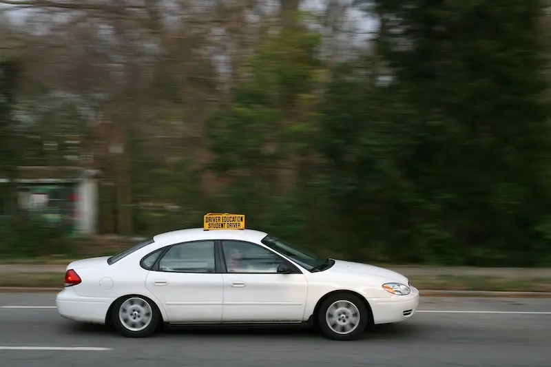 A white Ford Taurus driving down a road. It has a yellow Student Driver sign on top.